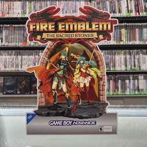 May include: A promotional display for the Game Boy Advance game "Fire Emblem: The Sacred Stones." The display features a colourful illustration of two characters with weapons, set against an archway. The game's title is prominently displayed at the top.