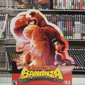May include: A promotional display for the Nintendo Switch game "Donkey Kong Bananza." The image features Donkey Kong and a young girl. The game's title is in yellow and orange, with the Nintendo Switch logo at the bottom. Shelves of video games are in the background.