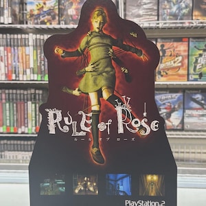 May include: A promotional display for the video game "Rule of Rose" for PlayStation 2. The display features a stylized image of a young woman in a green dress, with multiple arms reaching towards her. The game's title is prominently displayed.