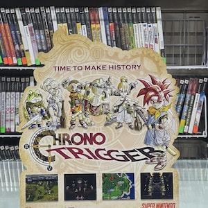Puede incluir: Un letrero promocional del juego de Super Nintendo "Chrono Trigger" con ilustraciones de personajes y el logotipo del juego. El letrero incluye el texto "Time to Make History" y capturas de pantalla del juego. El letrero se exhibe en una tienda de videojuegos.