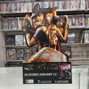 May include: A cardboard promotional display for the video game "Resident Evil 4" featuring character illustrations. The display includes the game title, release date, and the Nintendo GameCube logo. The background shows shelves of video games.
