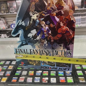 May include: A Final Fantasy Tactics PlayStation game display featuring a colorful illustration of characters. The display is approximately 6 inches wide. The game's title and the PlayStation and Square Soft logos are visible.