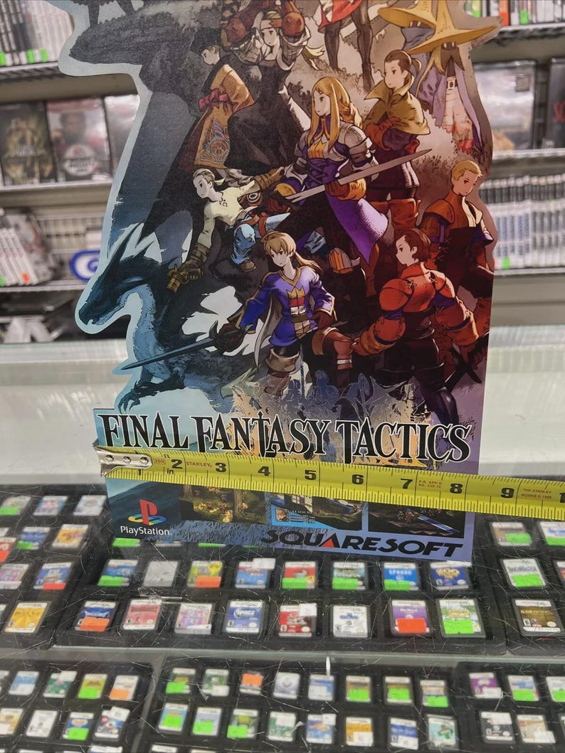 May include: A Final Fantasy Tactics PlayStation game display featuring a colorful illustration of characters. The display is approximately 6 inches wide. The game's title and the PlayStation and Square Soft logos are visible.