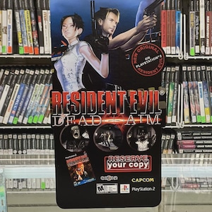 Puede incluir: Un letrero promocional del videojuego Resident Evil: Dead Aim, con dos personajes sosteniendo armas. El letrero incluye el título del juego en rojo y blanco, junto con el logotipo de PlayStation 2 y el texto "Reserva tu copia."