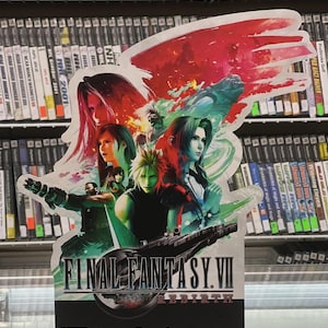 May include: A promotional sign for Final Fantasy VII Rebirth, featuring multiple characters and the game's title. The sign has a red and white design with the PS5, Epic Games Store, Steam, and Square Enix logos at the bottom. Shelves of video games form the background.