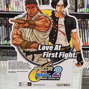 K&ouml;nnte beinhalten: Werbeschild f&uuml;r das Videospiel Capcom vs. SNK 2 mit Illustrationen von zwei Kampfspielcharakteren. Das Schild tr&auml;gt den Text "Love At First Fight" und Spieldetails. Das PlayStation 2-Logo ist sichtbar.