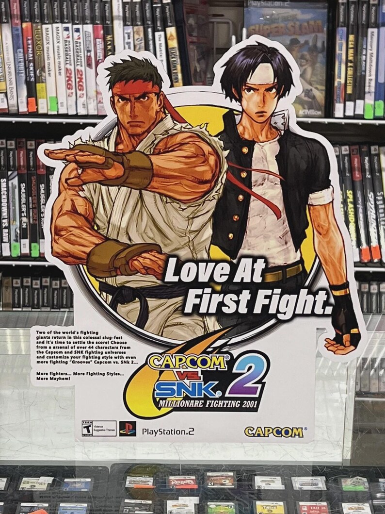 K&ouml;nnte beinhalten: Werbeschild f&uuml;r das Videospiel Capcom vs. SNK 2 mit Illustrationen von zwei Kampfspielcharakteren. Das Schild tr&auml;gt den Text "Love At First Fight" und Spieldetails. Das PlayStation 2-Logo ist sichtbar.