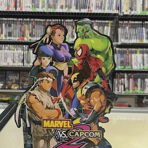 May include: A Marvel vs. Capcom 2 arcade game display featuring characters like Chun-Li, Hulk, Spider-Man, and Wolverine. The display has the game's logo and the text "New Age of Heroes" and "CAPCOM". The background shows a wall of video game cases.