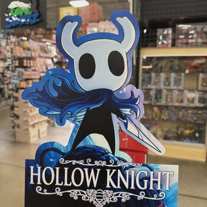 May include: A Hollow Knight promotional display featuring the character with a white head, large black eyes, and a blue cape. The display has the game's logo in white text on a blue background. The display is in a retail store.