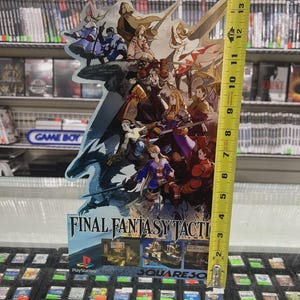 May include: A Final Fantasy Tactics promotional display, approximately 13 inches tall, featuring character artwork and the game's logo. The display is in front of a shelf of video game cartridges and a Game Boy sign.