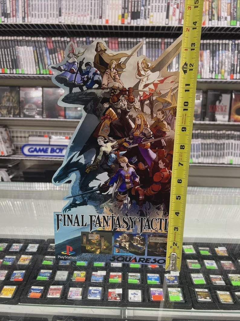 May include: A Final Fantasy Tactics promotional display, approximately 13 inches tall, featuring character artwork and the game's logo. The display is in front of a shelf of video game cartridges and a Game Boy sign.