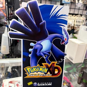 May include: A vertical rectangular display sign for the video game Pokémon XD: Gale of Darkness for the Nintendo GameCube. The sign features a large, dark blue Lugia creature with red eyes and the game title logo in yellow and red text.