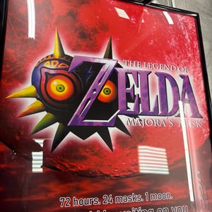 May include: A framed poster for The Legend of Zelda: Majora's Mask featuring the iconic purple and yellow mask against a vibrant red background. The text reads: 72 hours. 24 masks. 1 moon. The world is waiting on you, set in a black frame.