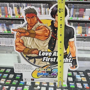 K&ouml;nnte beinhalten: Ein Papp-Werbedisplay f&uuml;r das Videospiel "Capcom vs. SNK 2: Millionaire Fighting 2001" mit Charakterillustrationen. Das Display ist etwa 33 cm hoch. Der Text "Love At First Fight" ist sichtbar.