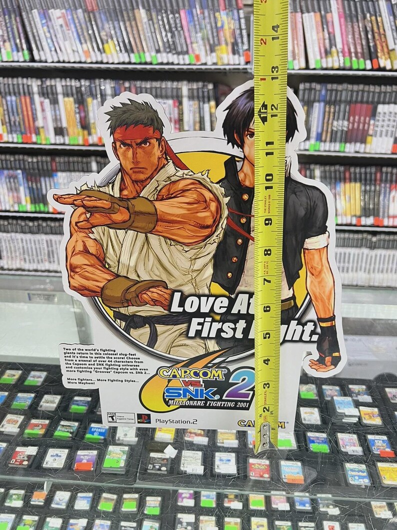 K&ouml;nnte beinhalten: Ein Papp-Werbedisplay f&uuml;r das Videospiel "Capcom vs. SNK 2: Millionaire Fighting 2001" mit Charakterillustrationen. Das Display ist etwa 33 cm hoch. Der Text "Love At First Fight" ist sichtbar.