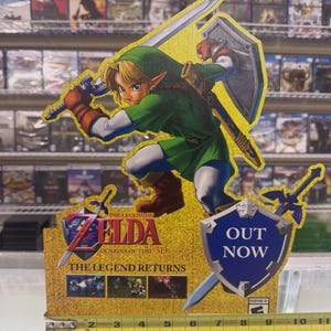 May include: A cardboard promotional display for The Legend of Zelda: Ocarina of Time 3D featuring Link with a sword and shield. The display includes the text 'OUT NOW' and 'THE LEGEND RETURNS' and measures approximately 26.7 cm wide at the base.