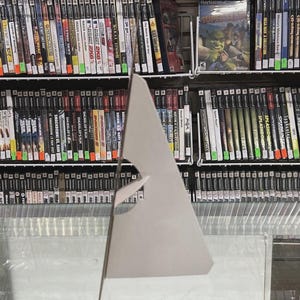 Puede incluir: Una exhibici&oacute;n de cajas de videojuegos en estanter&iacute;as. Las cajas son de varios tama&ntilde;os y colores, con t&iacute;tulos y gr&aacute;ficos visibles. Algunas cajas est&aacute;n etiquetadas como "PlayStation 2". El juego "Shrek" es visible. Debajo de las estanter&iacute;as hay cartuchos de juegos.