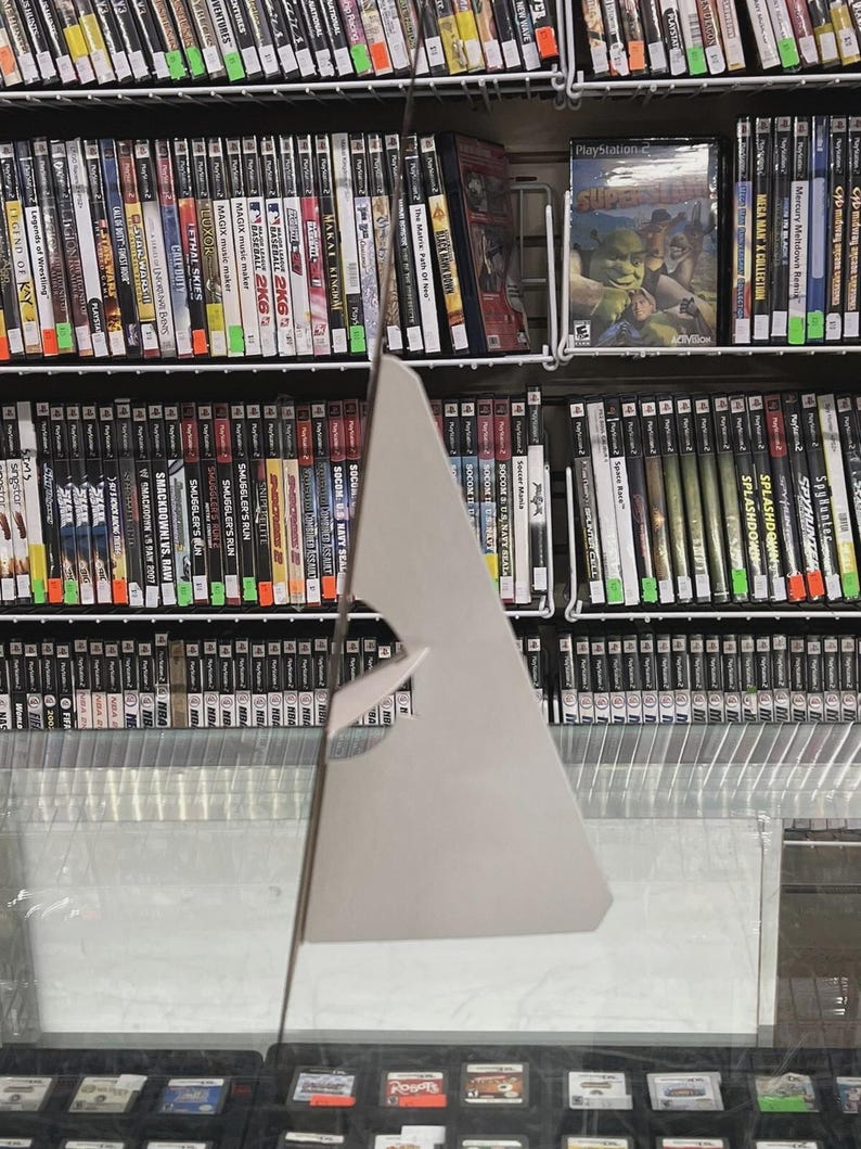 Puede incluir: Una exhibici&oacute;n de cajas de videojuegos en estanter&iacute;as. Las cajas son de varios tama&ntilde;os y colores, con t&iacute;tulos y gr&aacute;ficos visibles. Algunas cajas est&aacute;n etiquetadas como "PlayStation 2". El juego "Shrek" es visible. Debajo de las estanter&iacute;as hay cartuchos de juegos.