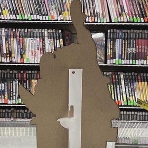Puede incluir: Un recorte de cart&oacute;n marr&oacute;n de una mano haciendo una se&ntilde;al de pulgar hacia arriba, con un soporte de pl&aacute;stico transparente adjunto en la parte posterior. El recorte est&aacute; frente a un estante lleno de cajas de videojuegos.