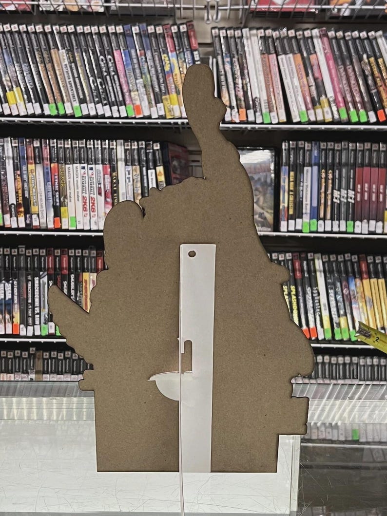 Puede incluir: Un recorte de cart&oacute;n marr&oacute;n de una mano haciendo una se&ntilde;al de pulgar hacia arriba, con un soporte de pl&aacute;stico transparente adjunto en la parte posterior. El recorte est&aacute; frente a un estante lleno de cajas de videojuegos.