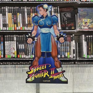 May include: A cardboard cutout of the Street Fighter II Turbo character Chun-Li, in a blue dress, stands in front of a shelf of video games. The cutout features the Capcom logo and the game's title. The background includes various game titles.