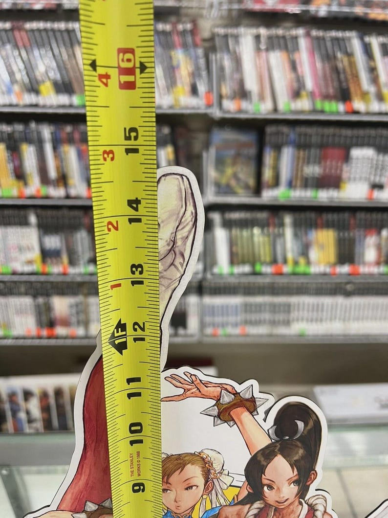 Puede incluir: Una imagen colorida y troquelada de personajes de videojuegos, de aproximadamente 30 cm de altura, se mide con una cinta m&eacute;trica amarilla. El fondo presenta estanter&iacute;as de carcasas de videojuegos. La imagen incluye el texto "THE STANLEY WORKS 1948".