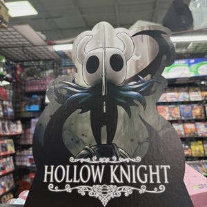 May include: A cardboard cutout featuring the Hollow Knight character from the video game. The cutout is primarily black and white, with the game's logo at the bottom. The character has a white head with horns and a long, pointed weapon.