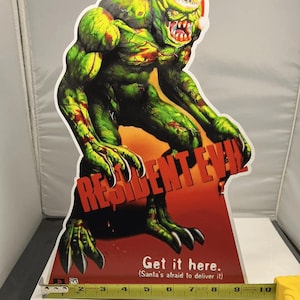 Puede incluir: Un recorte de cartón verde y rojo de Resident Evil de un monstruo con un gorro de Papá Noel. El recorte tiene el texto "RESIDENT EVIL" y "Get it here. (Santa's afraid to deliver it)". El recorte mide aproximadamente 23 cm de ancho.