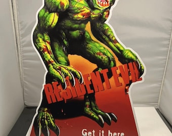 May include: A green and red Resident Evil cardboard cutout of a monster wearing a Santa hat. The cutout has the text "RESIDENT EVIL" and "Get it here. (Santa's afraid to deliver it)". The cutout is approximately 9 inches wide.