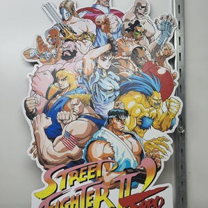 May include: A colorful Street Fighter II Turbo sign featuring multiple characters in dynamic poses. The sign has a white background with the game's logo in yellow, red, and pink. The characters are muscular and detailed.