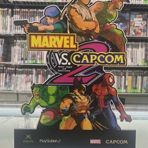 May include: A promotional sign for the video game "Marvel vs. Capcom 2: New Age of Heroes." The sign features illustrations of characters from both Marvel and Capcom, including Wolverine, Spider-Man, and Ryu. The sign also displays the logos for Xbox, PlayStation 2, Marvel, and Capcom.