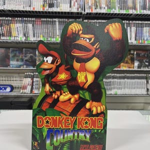 Może przedstawiać: Kartonowy wycinanka z Donkey Kong i Diddy Kong z gry wideo Donkey Kong Country. Obrazek przedstawia postacie stojące na beczce z logo gry na tle gier wideo.