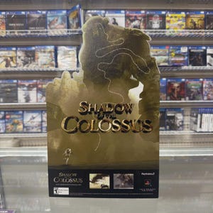 Può includere: Un espositore promozionale in cartone per il videogioco Shadow of the Colossus. L'espositore presenta una grande sagoma color marrone dorato di un colosso, il titolo del gioco in grassetto, una piccola figura e due immagini inserite con il marchio PlayStation 2.
