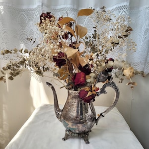 May include: A vintage silver teapot filled with dried flowers, including burgundy roses and tan foliage. The teapot has a decorative handle and spout, and sits on a small, ornate base. The arrangement is set against a white backdrop.
