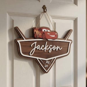Op de afbeelding: Houten deurbord met een rode cartoon raceauto en de naam "Jackson" in witte letters. Het bord heeft een bruin en wit schildontwerp met een geblokte vlag en het nummer "01".