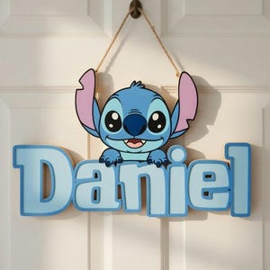 Può includere: Un'insegna in legno azzurro con il personaggio Stitch di Lilo & Stitch, con il nome "Daniel" in lettere azzurre. L'insegna è appesa a una porta con una corda marrone.