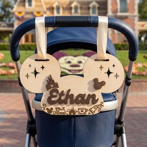 Può includere: Una targa in legno per passeggino a forma di testa di topo con il nome Ethan in corsivo marrone scuro. La superficie in legno chiaro presenta intagli di stelle, un castello e piccole icone tra cui un treno, una tazzina e un razzo, appesa con nastri color crema.