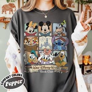 Puede incluir: Camiseta gris oscuro con un dise&ntilde;o de cuadr&iacute;cula que muestra personajes de Disney leyendo libros, como Mickey y Minnie Mouse. La camiseta incluye el texto "Walt Disney World Book Club" y el logotipo de Comfort Colors Pepper.