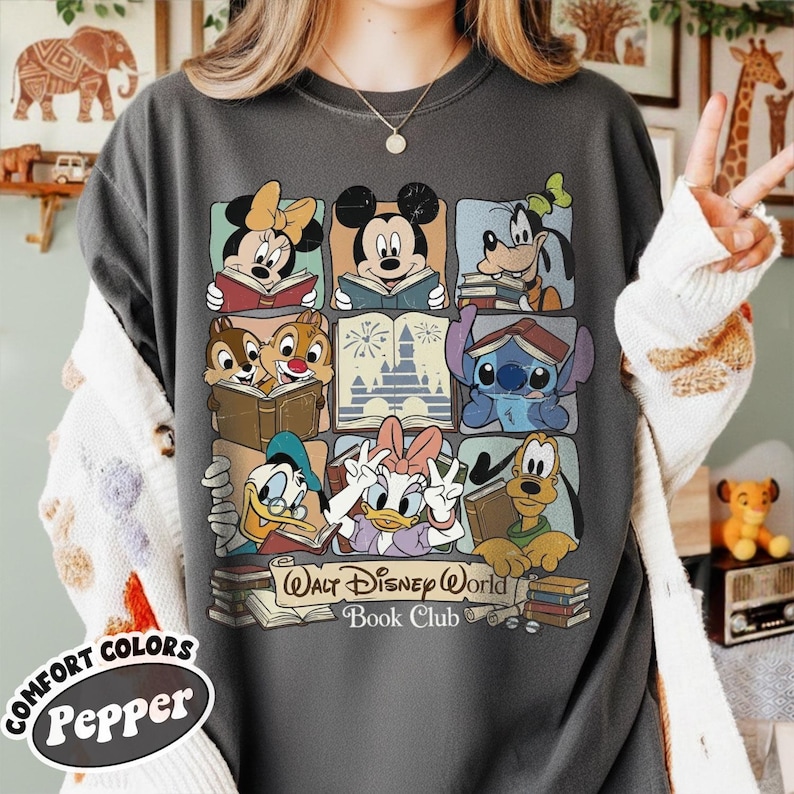 Puede incluir: Camiseta gris oscuro con un dise&ntilde;o de cuadr&iacute;cula que muestra personajes de Disney leyendo libros, como Mickey y Minnie Mouse. La camiseta incluye el texto "Walt Disney World Book Club" y el logotipo de Comfort Colors Pepper.
