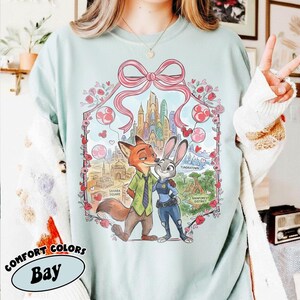 May include: A light blue Comfort Colors Bay t-shirt featuring a graphic of Nick Wilde and Judy Hopps from Zootopia. The design includes a pink bow, city skyline, and various districts. The shirt is a relaxed fit.