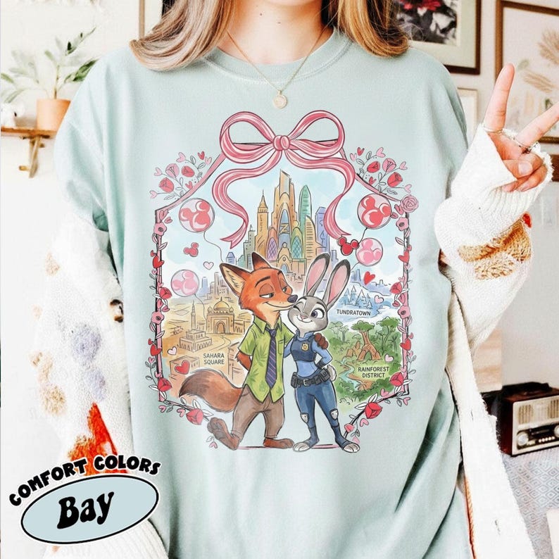 May include: A light blue Comfort Colors Bay t-shirt featuring a graphic of Nick Wilde and Judy Hopps from Zootopia. The design includes a pink bow, city skyline, and various districts. The shirt is a relaxed fit.