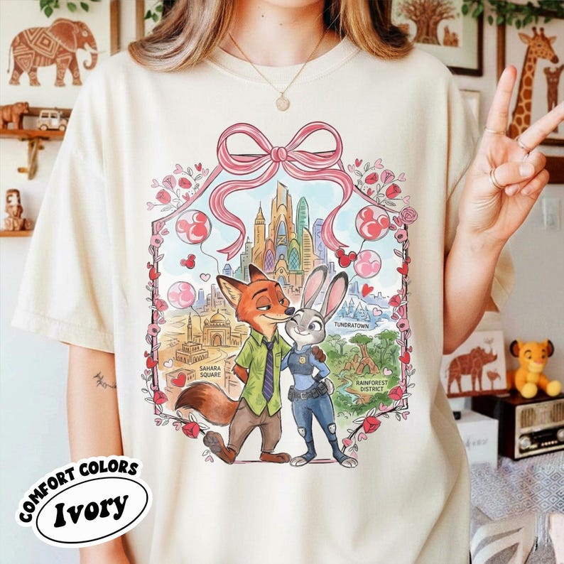 May include: Ivory-colored t-shirt featuring a colorful illustration of Nick Wilde and Judy Hopps from Zootopia, framed by a pink bow and floral elements. The shirt includes the text "COMFORT COLORS Ivory".