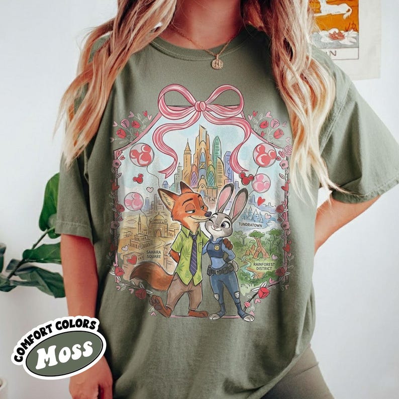 May include: Moss green t-shirt featuring a colorful graphic of Nick Wilde and Judy Hopps from Zootopia, with a cityscape and pink bow design. The shirt has the words "Comfort Colors" and "Moss" printed on it.