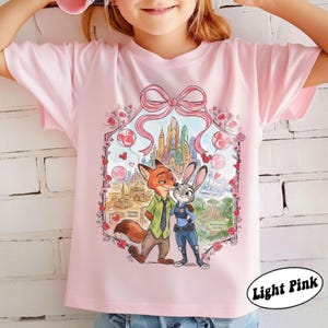 May include: Light pink t-shirt featuring a cartoon illustration of a fox and a rabbit embracing, with a cityscape backdrop. The design includes a pink bow and heart-shaped balloons. The text "Light Pink" is in a white oval.