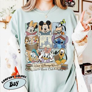 Puede incluir: Camiseta Comfort Colors azul claro con una cuadr&iacute;cula de personajes de Disney leyendo libros. La camiseta incluye a Mickey Mouse, Minnie Mouse, Goofy, el Pato Donald, Daisy, Chip y Chop, Stitch y Pluto. El texto "Walt Disney World Book Club" tambi&eacute;n est&aacute; impreso.