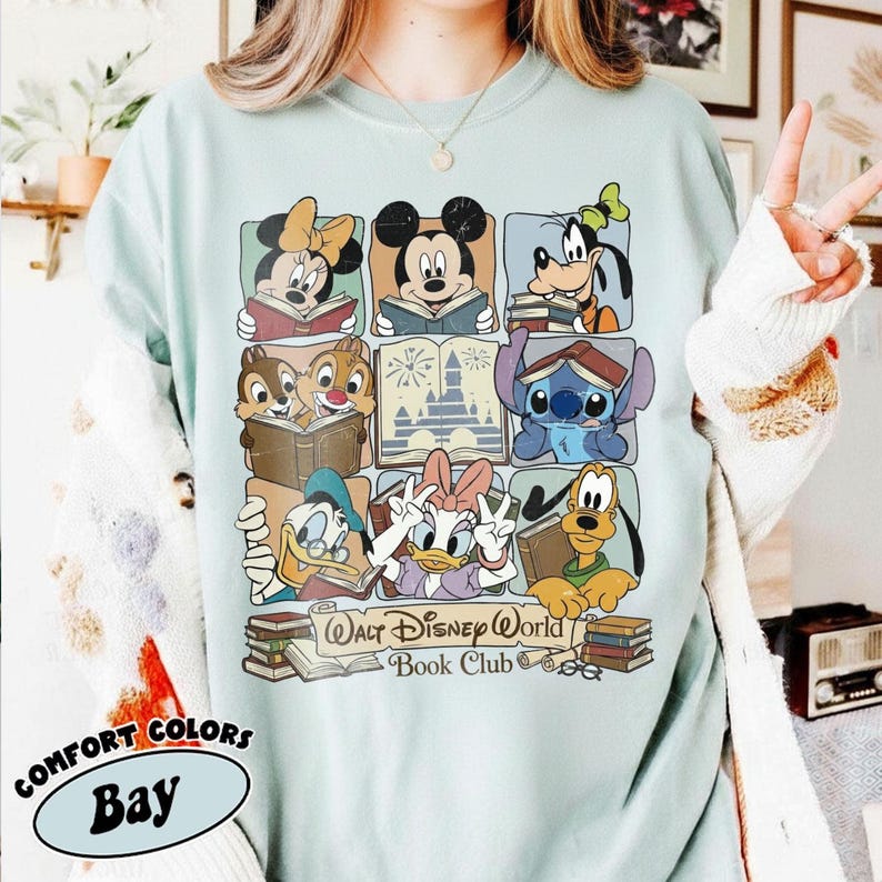 Puede incluir: Camiseta Comfort Colors azul claro con una cuadr&iacute;cula de personajes de Disney leyendo libros. La camiseta incluye a Mickey Mouse, Minnie Mouse, Goofy, el Pato Donald, Daisy, Chip y Chop, Stitch y Pluto. El texto "Walt Disney World Book Club" tambi&eacute;n est&aacute; impreso.