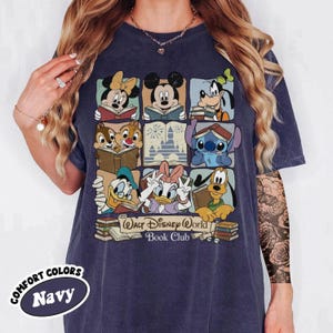 Puede incluir: Camiseta azul marino con un dise&ntilde;o de cuadr&iacute;cula de personajes de Disney leyendo libros, incluyendo Mickey y Minnie Mouse. La camiseta tiene el texto "Walt Disney World Book Club" y "Comfort Colors" impresos.
