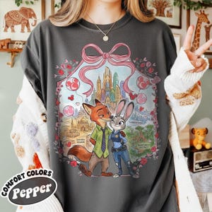 May include: Dark gray t-shirt featuring a cartoon illustration of Nick Wilde and Judy Hopps from Zootopia, framed by a pink bow and floral elements. The shirt has the words "COMFORT COLORS Pepper" printed on the bottom left.