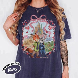 May include: Navy blue t-shirt featuring a colorful illustration of Nick Wilde and Judy Hopps from Zootopia. The design includes a cityscape, hearts, and the words "Sahara Square", "Tundratown", and "Rainforest District". The shirt has the words "Comfort Colors" and "Navy".