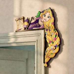 May include: A decorative wooden wall hanging featuring a cartoon image of a princess with long blonde hair adorned with flowers. The princess is wearing a purple dress and has a green chameleon on her shoulder. A palette and paintbrush are also included.